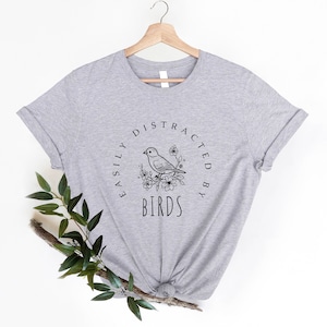 K&ouml;nnte beinhalten: Graues T-Shirt mit einer schwarz-wei&szlig;en Illustration eines Vogels auf einem Ast mit Blumen. Der Text "Easily Distracted By Birds" ist in einem Kreis um den Vogel geschrieben.