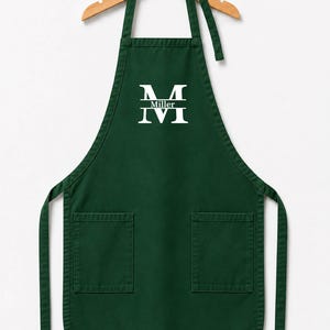 Puede incluir: Un delantal verde oscuro con dos bolsillos delanteros y un estampado gráfico blanco en el pecho. El estampado muestra una letra M grande, la palabra Miller y la letra M debajo. El delantal cuelga de una percha de madera con una pequeña bola naranja.