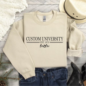 Könnte beinhalten: Ein beiger Pullover mit dem Aufdruck "CUSTOM UNIVERSITY EST. 1973 mom" auf der Vorderseite.