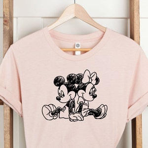 Puede incluir: Una camiseta rosa con una ilustraci&oacute;n en blanco y negro de Mickey Mouse y Minnie Mouse cogidos de la mano.