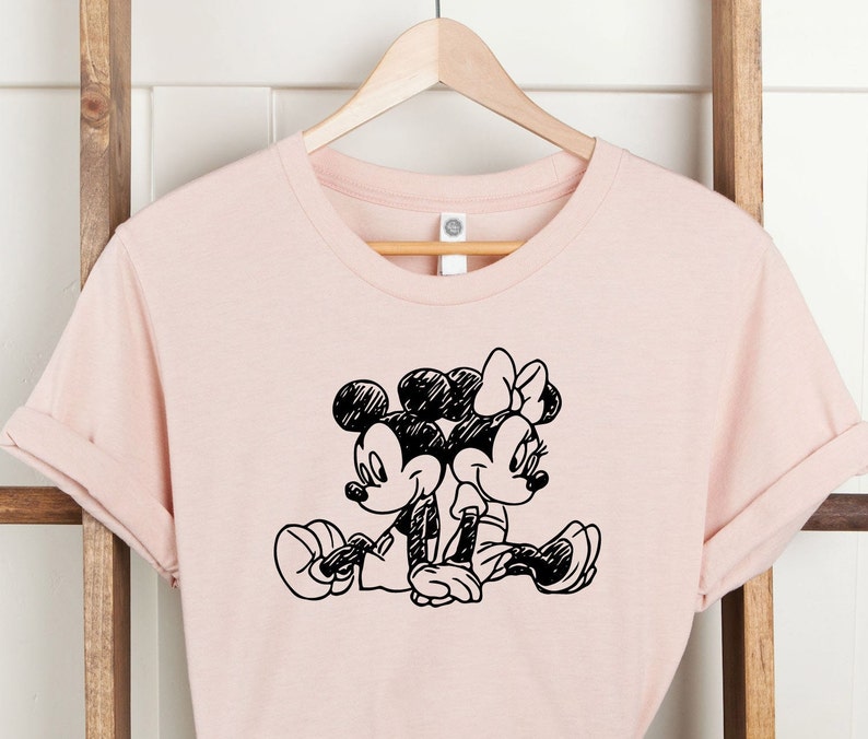 Puede incluir: Una camiseta rosa con una ilustraci&oacute;n en blanco y negro de Mickey Mouse y Minnie Mouse cogidos de la mano.
