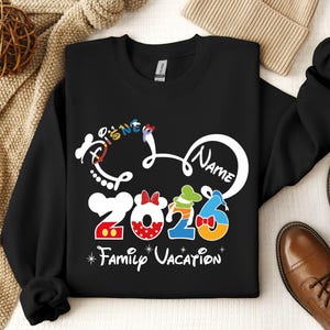 A black sweatshirt with a Disney-themed design, including the text "Disney Name" and "2026 Family Vacation", along with various Disney characters and numbers. The sweatshirt is placed on a surface with a beige blanket and a pair of brown shoes.
