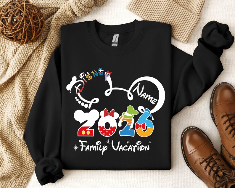 A black sweatshirt with a Disney-themed design, including the text "Disney Name" and "2026 Family Vacation", along with various Disney characters and numbers. The sweatshirt is placed on a surface with a beige blanket and a pair of brown shoes.