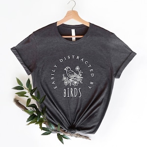 K&ouml;nnte beinhalten: Ein dunkelgraues T-Shirt mit einem wei&szlig;en Grafikdesign, auf dem "Easily Distracted By Birds" mit einer stilisierten Vogel-Illustration steht.