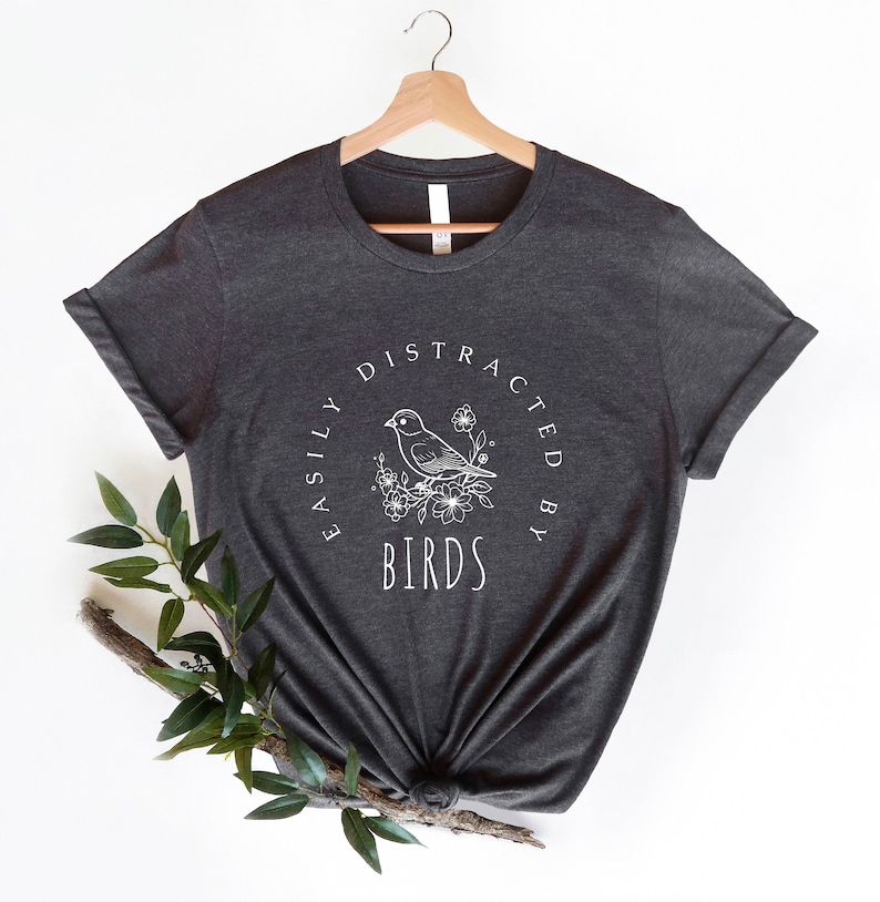 K&ouml;nnte beinhalten: Ein dunkelgraues T-Shirt mit einem wei&szlig;en Grafikdesign, auf dem "Easily Distracted By Birds" mit einer stilisierten Vogel-Illustration steht.