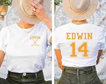 May include: White t-shirt with a gold embroidered hockey stick design and the text "EDWIN 14" on the back.