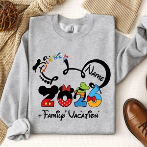 A gray sweatshirt with a Disney-themed design, including the text "Disney Name" and "2016 Family Vacation", along with various Disney characters and numbers. The sweatshirt is placed on a surface with other items such as a beige blanket, a pair of brown shoes, and a woven basket.