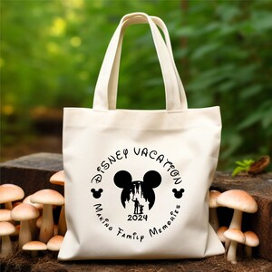 Peut inclure: Sac fourre-tout en toile blanche avec un motif circulaire noir repr&eacute;sentant les oreilles de Mickey Mouse, une silhouette du ch&acirc;teau de Cendrillon et le texte "Disney Vacation Making Family Memories 2024".