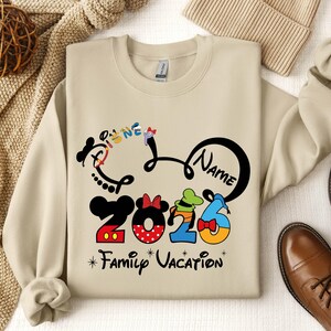 A beige sweatshirt with a Disney-themed design, including a Mickey Mouse and Minnie Mouse logo, along with the text "Disney 2026 Family Vacation". The sweatshirt is laid out on a surface with other clothing items and accessories, such as a sweater, a hat, and a pair of shoes.
