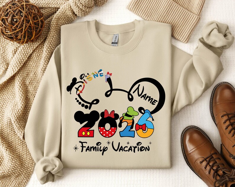 A beige sweatshirt with a Disney-themed design, including a Mickey Mouse and Minnie Mouse logo, along with the text "Disney 2026 Family Vacation". The sweatshirt is laid out on a surface with other clothing items and accessories, such as a sweater, a hat, and a pair of shoes.
