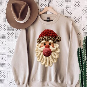 Peut inclure: Sweat-shirt beige avec un motif de Père Noël. Le Père Noël a un bonnet rouge tricoté avec un bord léopard, un nez rose et une barbe en fil blanc. Un chapeau marron avec une bande blanche est suspendu à gauche.