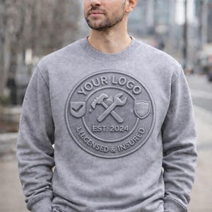 May include: A gray sweatshirt with a circular logo featuring a hammer, wrench, and shield. The logo includes the text "YOUR LOGO", "EST. 2024", and "LICENSED & INSURED". The sweatshirt is a solid gray color.