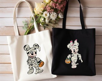 Könnte beinhalten: Zwei Halloween-Tragetaschen. Eine ist cremefarben mit einer Mumie-Mickey-Mouse-Grafik, die einen Kürbis hält. Die andere ist schwarz mit einer Mumie-Minnie-Mouse-Grafik, die einen Kürbis hält. Beide Taschen haben lange Henkel.