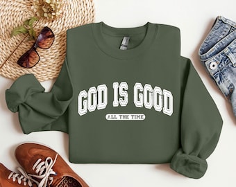 May include: Olive green sweatshirt with the text "GOD IS GOOD ALL THE TIME" in white, arched lettering. The sweatshirt is paired with blue jeans, brown shoes, and sunglasses.