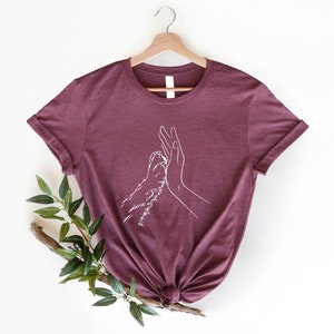 May include: Burgundy t-shirt with a white line drawing of a human hand and a paw giving a high five. The shirt is hanging on a wooden hanger. A sprig of greenery is in the lower left corner.