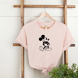 May include: A pink t-shirt with a black silhouette of Mickey Mouse walking. The t-shirt is hanging on a wooden ladder.