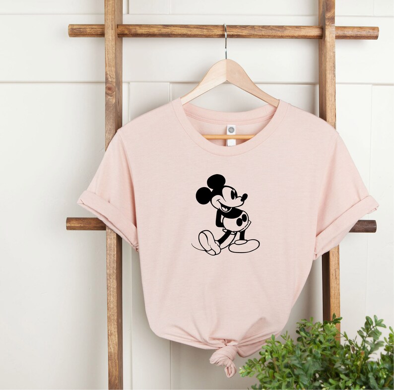 May include: A pink t-shirt with a black silhouette of Mickey Mouse walking. The t-shirt is hanging on a wooden ladder.