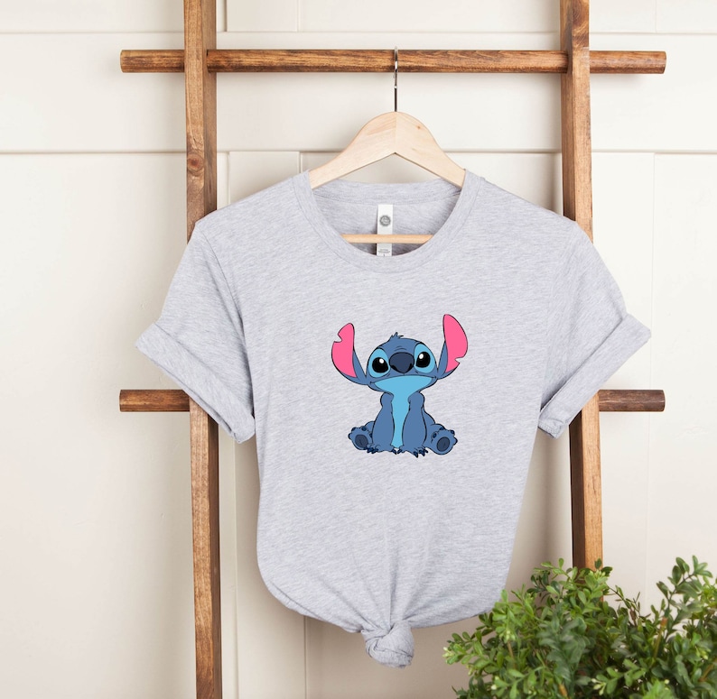 May include: A gray t-shirt with a blue and pink cartoon character of Stitch from the Disney movie Lilo & Stitch. The character is sitting with its arms crossed and has a big smile.