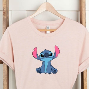 May include: A pink t-shirt with a blue Stitch character from Lilo & Stitch. Stitch is sitting with his arms crossed and has a big smile on his face.
