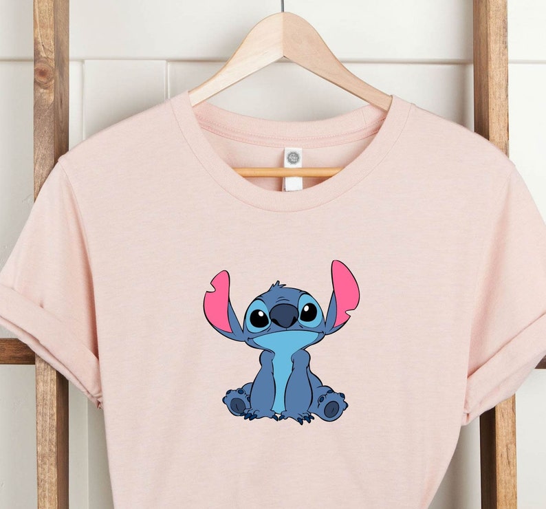 May include: A pink t-shirt with a blue Stitch character from Lilo & Stitch. Stitch is sitting with his arms crossed and has a big smile on his face.