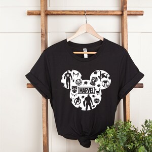 May include: Black t-shirt with a white Mickey Mouse silhouette design featuring Marvel characters, including Iron Man, Captain America, Thor, Black Panther, and more. The word "MARVEL" is printed inside the silhouette.