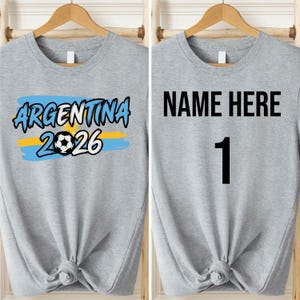 May include: Two light gray t-shirts hanging on wooden hangers. One shirt features the text "ARGENTINA 2026" with a soccer ball graphic. The other shirt has "NAME HERE" and the number "1" printed in black.