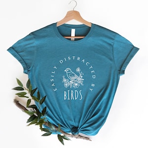 K&ouml;nnte beinhalten: Tealblaues T-Shirt mit einer wei&szlig;en Grafik eines Vogels auf einem Ast mit Blumen und dem Text "Easily Distracted By Birds".