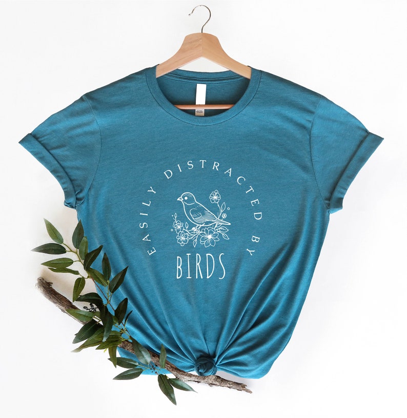 K&ouml;nnte beinhalten: Tealblaues T-Shirt mit einer wei&szlig;en Grafik eines Vogels auf einem Ast mit Blumen und dem Text "Easily Distracted By Birds".