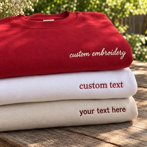 May include: A stack of three crewneck sweatshirts in red, white, and beige. The red sweatshirt features white cursive text reading custom embroidery. The white sweatshirt has red text reading custom text. The beige sweatshirt has red text reading your text here.