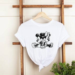 Puede incluir: Camiseta blanca con un boceto en blanco y negro de Mickey Mouse y Minnie Mouse sentados juntos.
