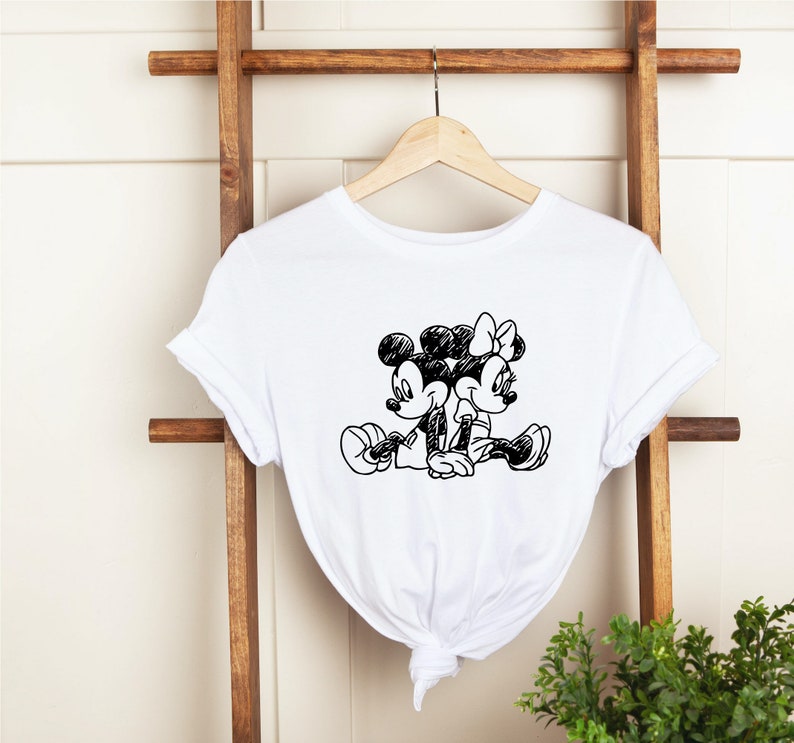 Puede incluir: Camiseta blanca con un boceto en blanco y negro de Mickey Mouse y Minnie Mouse sentados juntos.