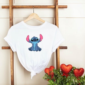 May include: White t-shirt with a blue Stitch character from the Disney movie Lilo & Stitch. Stitch is sitting with his arms crossed and has pink ears.