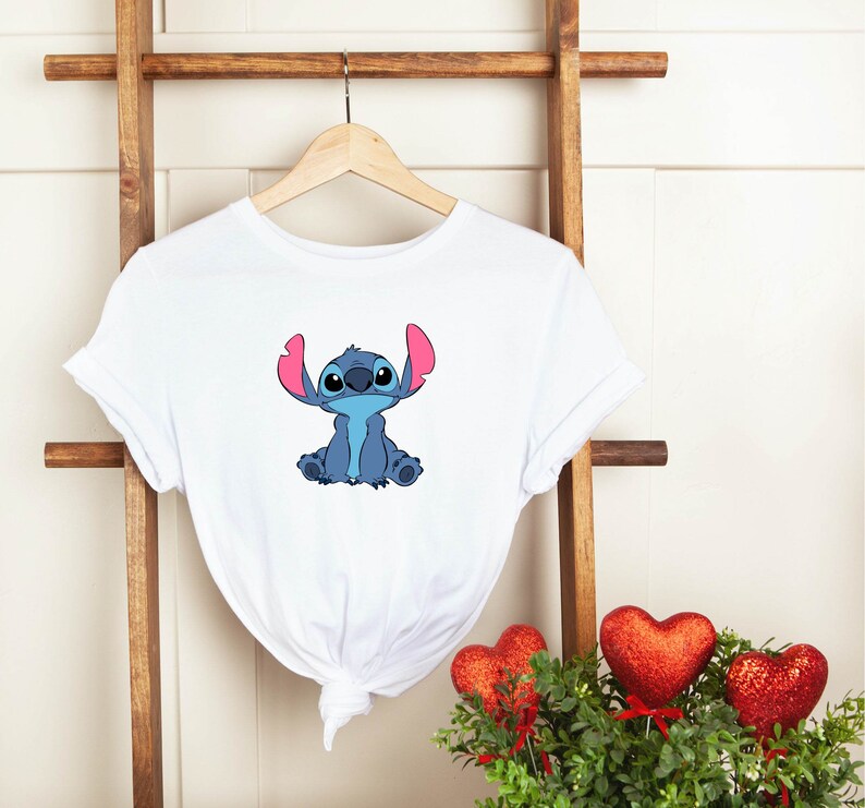 May include: White t-shirt with a blue Stitch character from the Disney movie Lilo & Stitch. Stitch is sitting with his arms crossed and has pink ears.