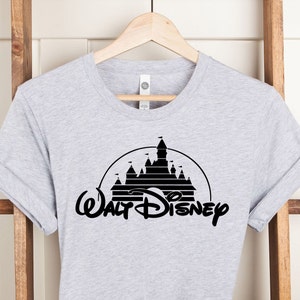 K&ouml;nnte beinhalten: Hellgraues T-Shirt mit einem schwarzen Grafikdesign eines Schlosses in einer Halbkreisform, darunter die Worte "Walt Disney" in einer stilisierten Schriftart. Das T-Shirt h&auml;ngt an einem Holzkleiderb&uuml;gel vor einem wei&szlig;en Hintergrund.