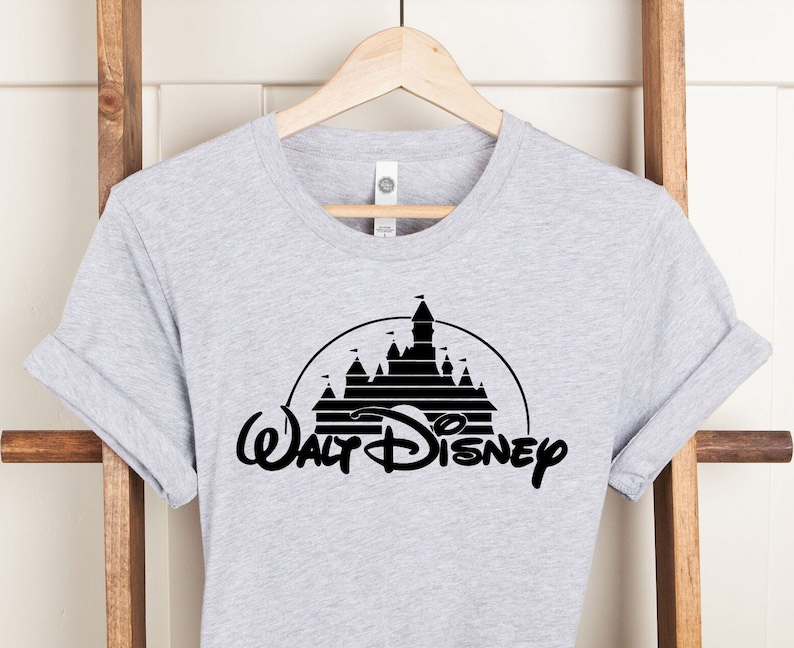 K&ouml;nnte beinhalten: Hellgraues T-Shirt mit einem schwarzen Grafikdesign eines Schlosses in einer Halbkreisform, darunter die Worte "Walt Disney" in einer stilisierten Schriftart. Das T-Shirt h&auml;ngt an einem Holzkleiderb&uuml;gel vor einem wei&szlig;en Hintergrund.