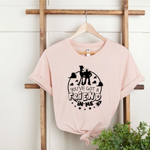 Może przedstawiać: R&oacute;żowy T-shirt z czarną sylwetką Woody'ego i Buzz Lightyeara z Toy Story. Na koszulce nadrukowany jest tekst "You've got a friend in me".