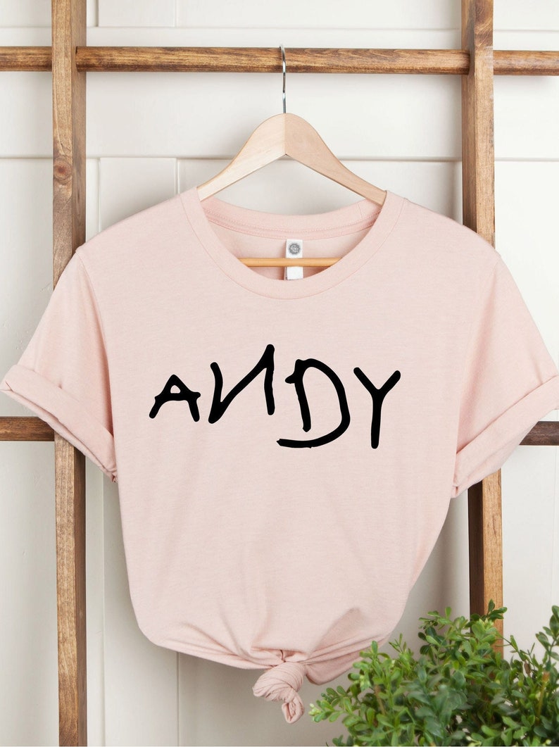 Może przedstawiać: R&oacute;żowa koszulka z napisem "ANDY" w kolorze czarnym na przodzie. Koszulka jest na wieszaku.