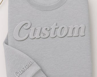 Puede incluir: Sudadera gris claro con la palabra "Custom" en relieve en la parte delantera y en la manga. La sudadera está doblada cuidadosamente, con una etiqueta en el cuello. Una flor de diente de león está en la esquina superior izquierda.