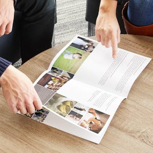 May include: An open brochure on a wooden table, featuring wedding-themed images and text. The brochure is being held open by two people, with one person pointing at the text. The brochure has multiple images of weddings.