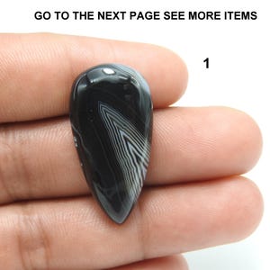 Pode incluir: Uma pedra preciosa em forma de gota, polida, preta e branca, segurada na m&atilde;o. A pedra apresenta um padr&atilde;o listrado. O texto "GO TO THE NEXT PAGE SEE MORE ITEMS" e o n&uacute;mero "1" s&atilde;o vis&iacute;veis.