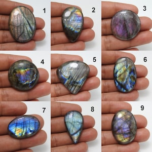 May include: Nine polished labradorite gemstones in various shapes, including oval, teardrop, and round. The stones display iridescent flashes of blue, purple, and gold within a gray base. Each stone is held in a hand, showcasing the natural beauty and unique patterns.