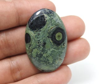 May include: An oval-shaped gemstone with a unique pattern. The stone features a mottled green base with black, circular inclusions resembling eyes. The polished surface reflects light, highlighting the natural variations in color and texture. A striking piece for jewelry or collection.