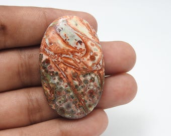 May include: An oval-shaped gemstone with a unique pattern of red, brown, green, and cream colors. The polished surface displays a marbled effect, with intricate veining and speckled textures. The stone is held in a hand against a white background.