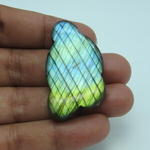 Peut inclure: Une pierre précieuse de labradorite polie, de forme organique unique. La pierre présente un jeu de couleurs vibrant, avec des teintes de bleu, de vert et de jaune. La surface présente un motif subtil en forme de grille. La pierre est tenue dans une main.