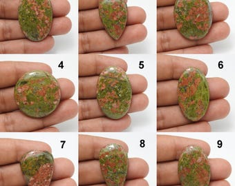 May include: Nine polished unakite gemstones in various shapes, including oval, teardrop, and round. The stones display a mix of green and pink hues, with a speckled pattern. Each stone is held in a hand, showcasing the natural variations in color and form.