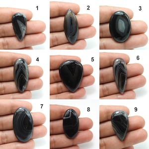Pode incluir: Nove pedras preciosas de &aacute;gata polida em preto e branco em v&aacute;rias formas, incluindo ovais, em forma de gota e alongadas. Cada pedra exibe padr&otilde;es de bandas &uacute;nicos, com um acabamento brilhante. As pedras preciosas s&atilde;o seguradas em uma m&atilde;o, mostrando seu tamanho e detalhes.