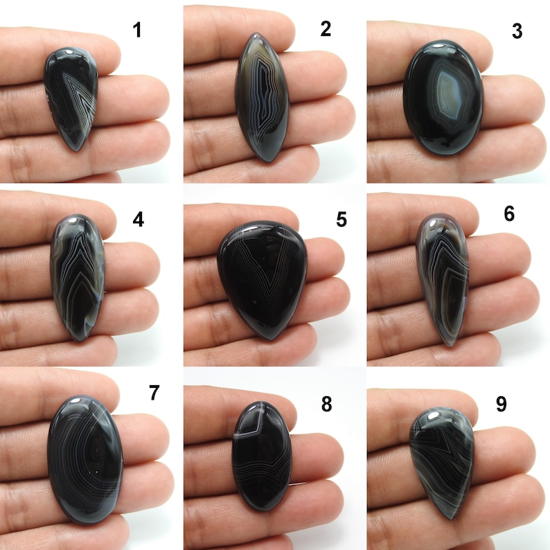 Pode incluir: Nove pedras preciosas de &aacute;gata polida em preto e branco em v&aacute;rias formas, incluindo ovais, em forma de gota e alongadas. Cada pedra exibe padr&otilde;es de bandas &uacute;nicos, com um acabamento brilhante. As pedras preciosas s&atilde;o seguradas em uma m&atilde;o, mostrando seu tamanho e detalhes.