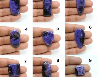 May include: A collection of nine polished gemstones in various shapes, including oval, rectangular, and teardrop. The stones feature a striking combination of deep purple and black hues, with some displaying a marbled effect. Each stone is held in a hand against a white background.