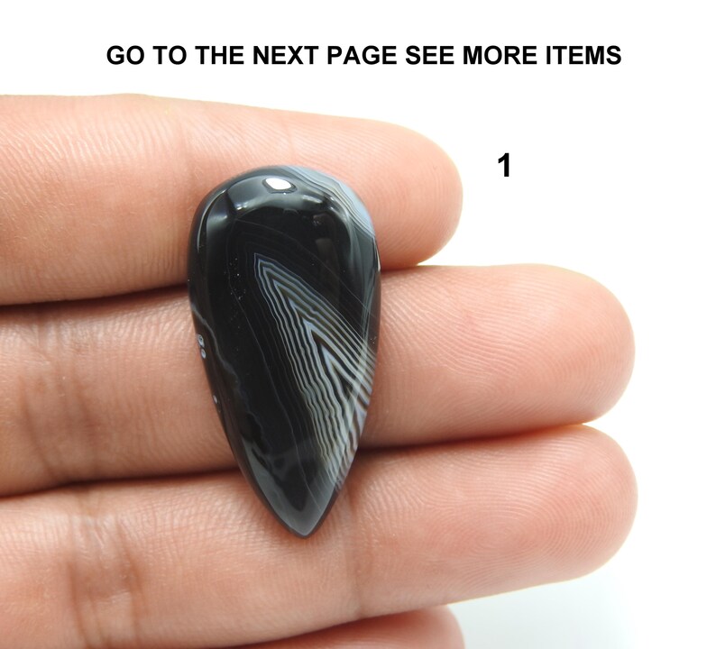 Pode incluir: Uma pedra preciosa em forma de gota, polida, preta e branca, segurada na m&atilde;o. A pedra apresenta um padr&atilde;o listrado. O texto "GO TO THE NEXT PAGE SEE MORE ITEMS" e o n&uacute;mero "1" s&atilde;o vis&iacute;veis.