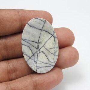 May include: Oval-shaped gemstone with a light gray and green base, crisscrossed by dark gray veins. The stone is held in a hand, showcasing its polished surface and natural pattern. A unique piece for jewelry or collection.